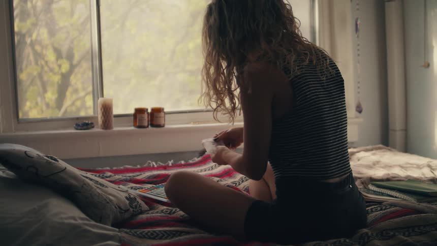 A woman sitting on a bed with a blanket and pillow with a window in the background