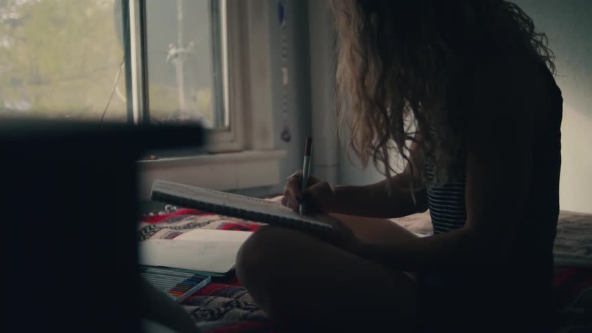 A young girl with curly hair sits cross legged on her bed as she does homework