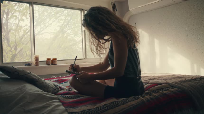 Blonde woman with curly hair sitting on a bed writes things in a notebook in her room