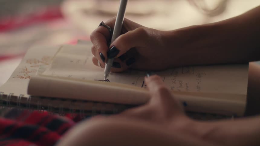 Woman sitting on bed draws something in the notebook with a pen