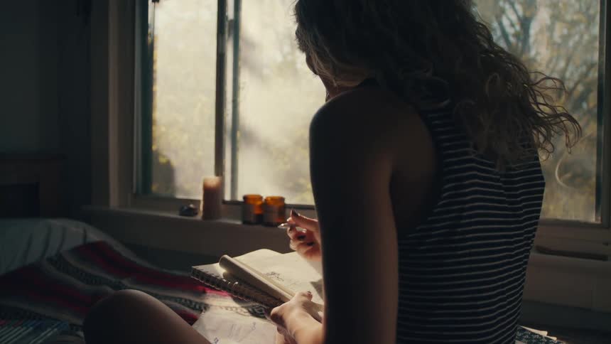 Sunlights shines onto a woman as she draws in a notebook while sitting upright in a bed next to a window