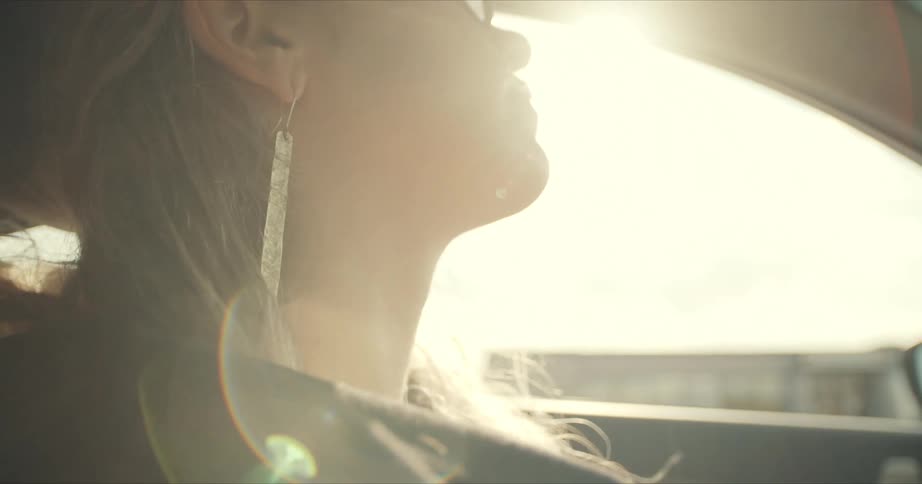 Side view of a blonde woman with earrings driving a car as the sun shines brightly in the background