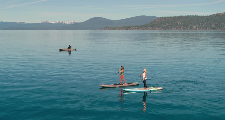 Two women on a lake paddle boarding with a person on a boat a mountain range and the sky in the background