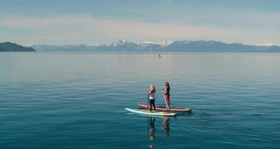 Two women paddle boarding in the water with boats and mountains in the distance