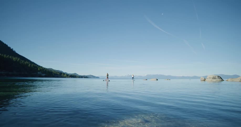 Two women stand on paddleboards paddle through a turquoise tranquil lake against blue sky