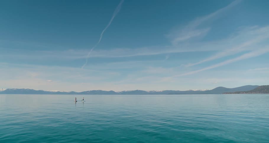 Aerial view of two women standing on paddle boards paddle through a turquoise tranquil lake against blue sky