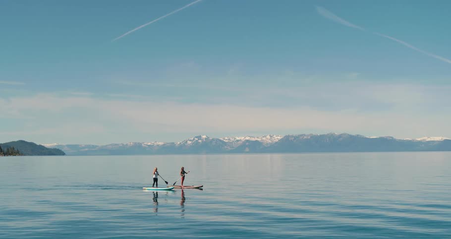 A pair of women standing on paddleboards paddle side by side across a lake surrounded by mountains on a sunny day