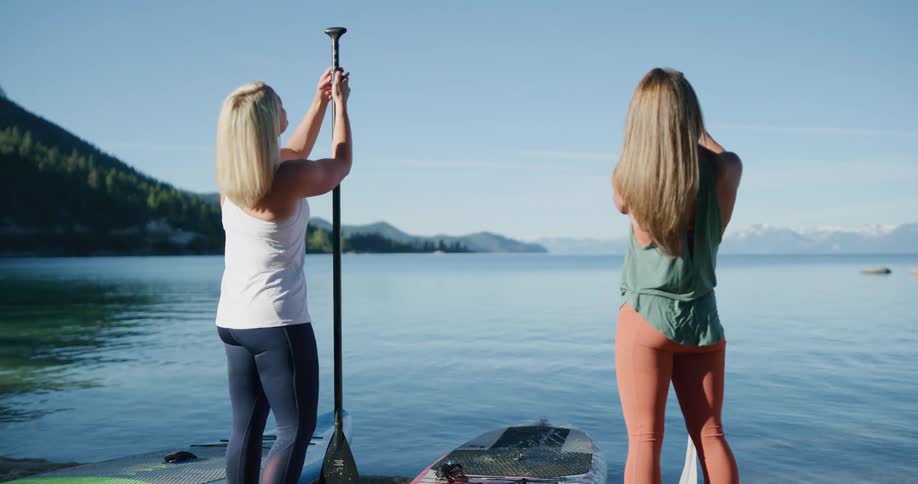 A pair of women standing on the shore of a lake adjust their paddles as they prepare to paddle board across the lake