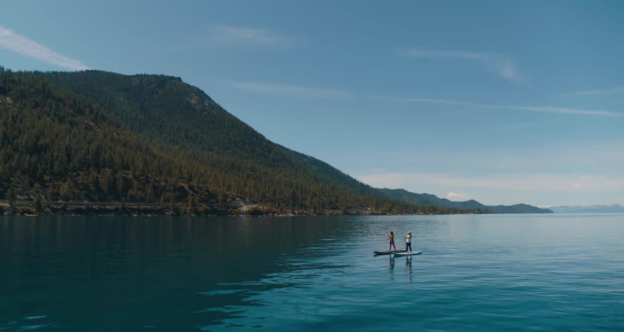 Two women standing on paddle boards paddle through a tranquil lake with green mountains in the background