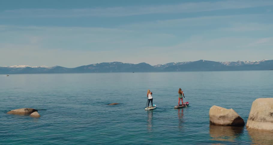 Aerial view of two women standing on paddle boards paddling through a tranquil lake with mountains in the background