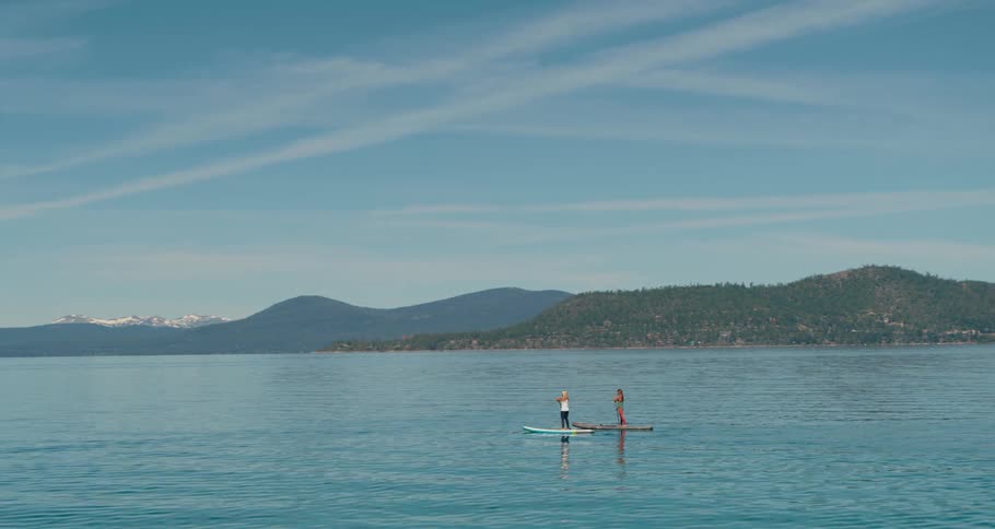 Two women paddle board across a large body of water near forested hills