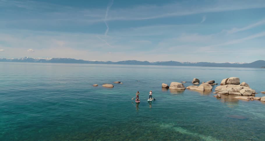 Aerial view of two women standing on paddle boards paddle through a turquoise tranquil lake against blue sky