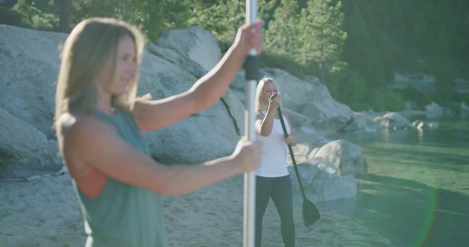 Two women twist together long oars to use with their paddle boards on a lake