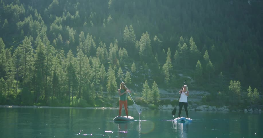 Two women standing on paddle boards paddle through a tranquil lake with green mountains in the background