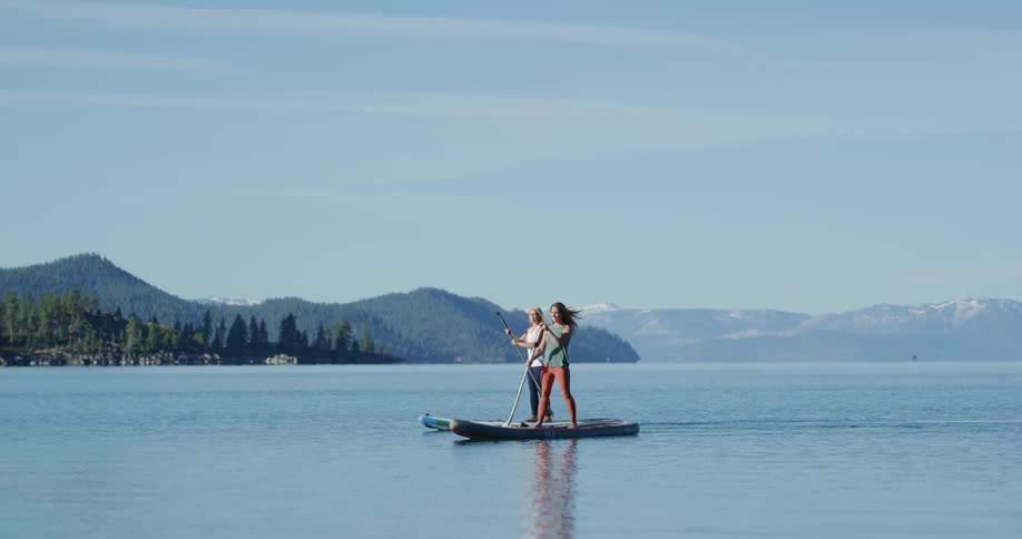 Two women paddle boarding on a lake with a mountain range and the sky in the background