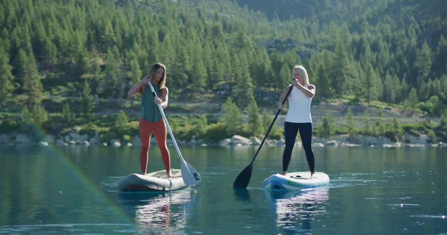 Two women standing on paddleboards paddle through a tranquil lake with green mountains in the background