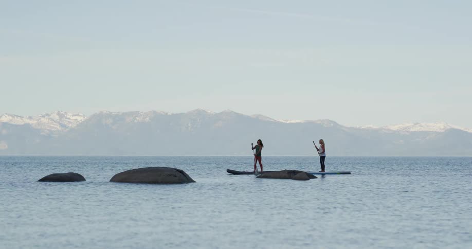 Two women paddle boarding on a lake with a mountain range and the sky in the background