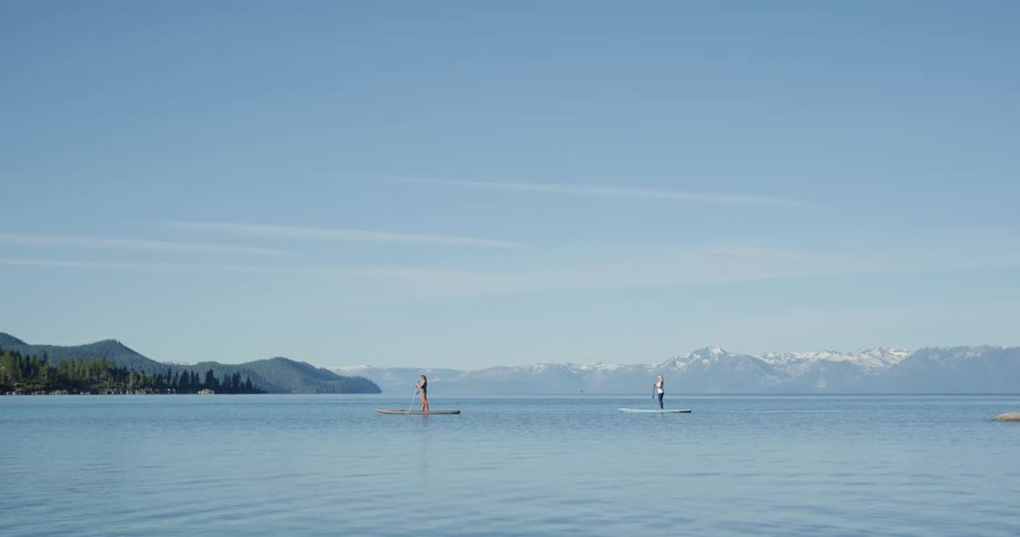 A pair of women cross a lake on paddleboards on a sunny day