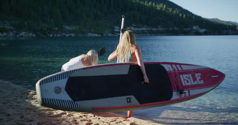 Two fit woman carry paddle boards to the shore of a lake in the mountains