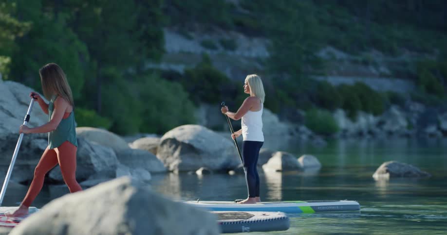 Two fit women paddle boarding over a lake in the mountains at morning