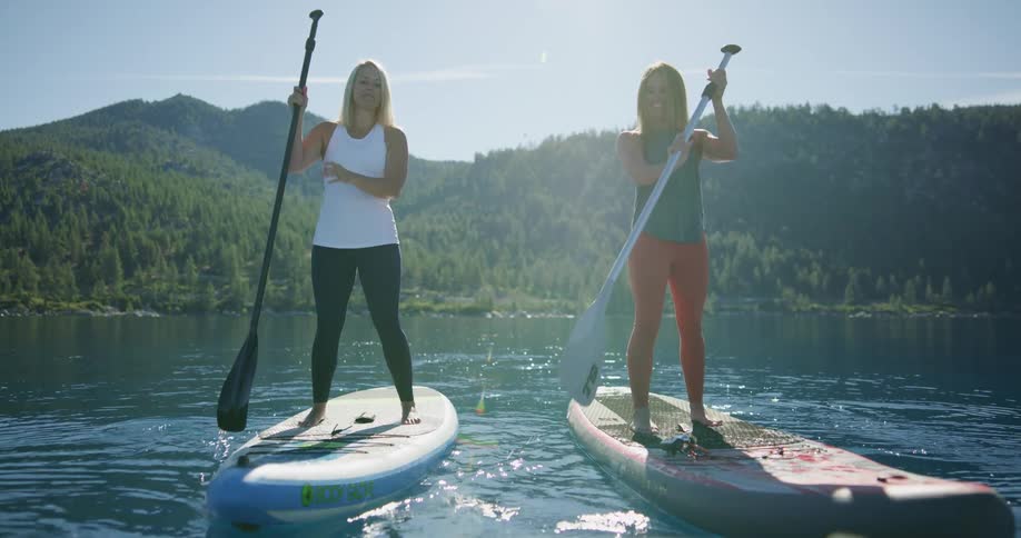 Two women standing on paddleboards paddle through a tranquil lake with green mountains in the background