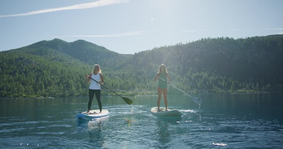 Two women standing on paddle boards paddle through a tranquil lake with green mountains in the background