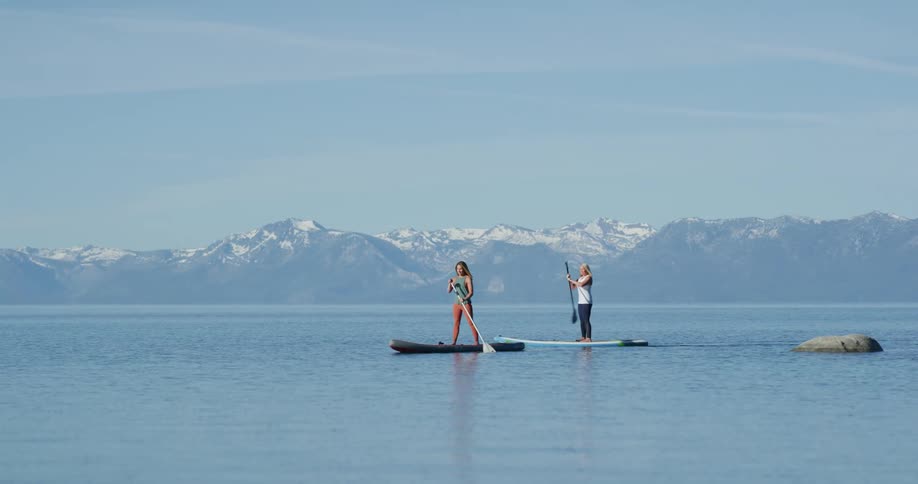 Two women paddle boarding on a lake with the mountains in the distance