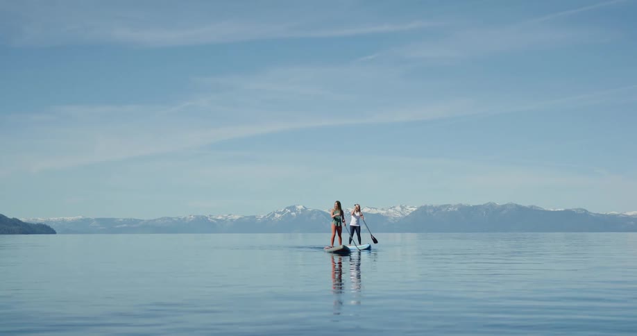 Two women paddle boarding on a lake with a mountain range and the sky in the background