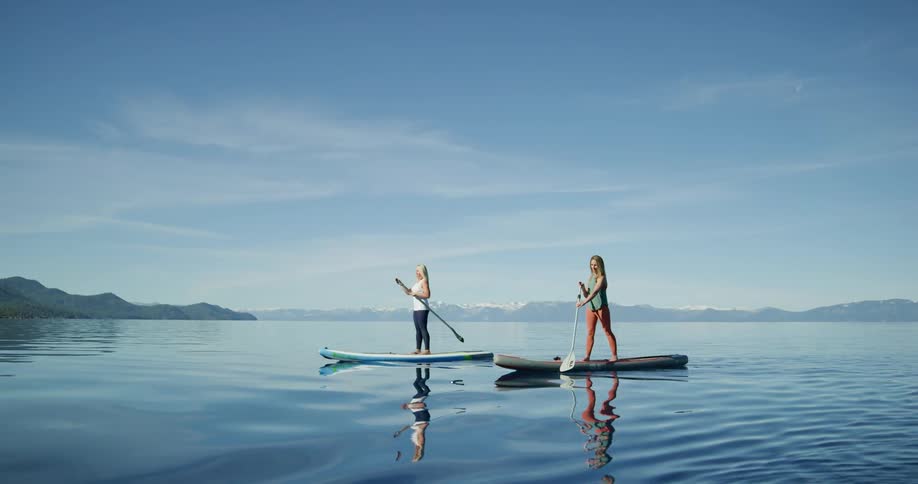 Two women standing on paddle boards paddle through a tranquil lake against the blue sky