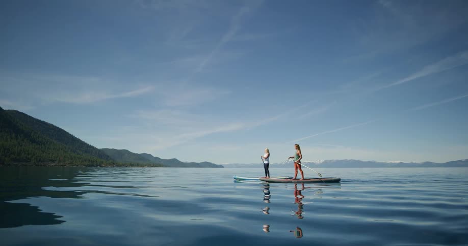 A pair of women stand on paddle boards and use their paddles to travel side by side towards the shore