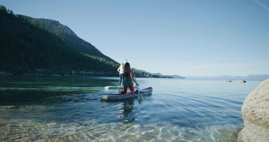 Two fit woman paddle on boards over the surface of a lake in the mountains