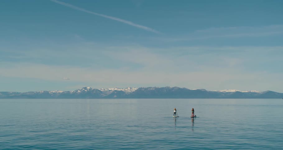 Two women paddle boarding on a lake with a mountain range and the sky in the background