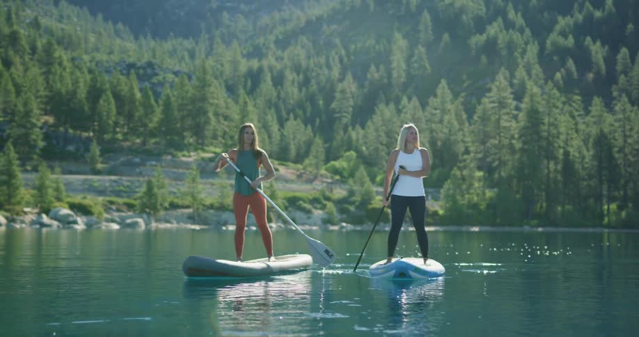 Two women uses oars to push standing paddle boats along a river