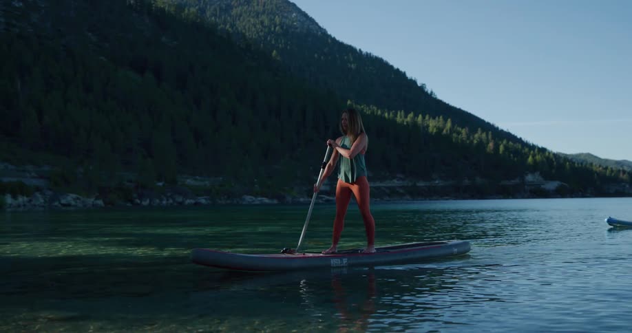 Two women standing on paddle boards paddle through a tranquil lake and arrives on land with mountains in the background