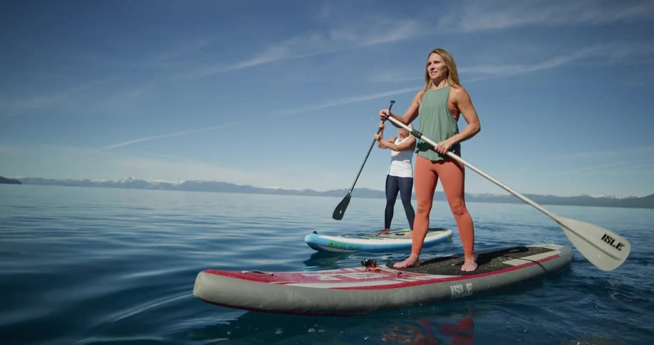 A pair of women cross a lake on paddle boards on a sunny day