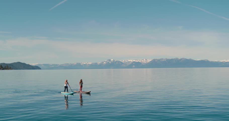 Two women on a lake paddleboarding with a mountain range and the sky in the background