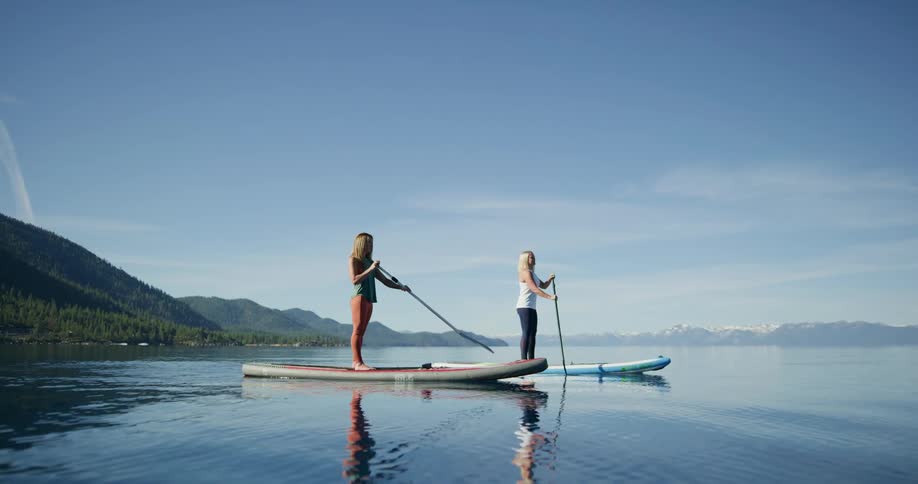 Two woman paddle board along a large lake