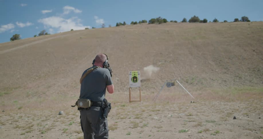 A man wearing hearing protection aims and shoots a gun at a paper target placed alongside a hill