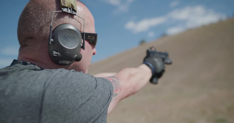 A man with gloves earmuffs and glasses aiming and shooting a pistol at a target