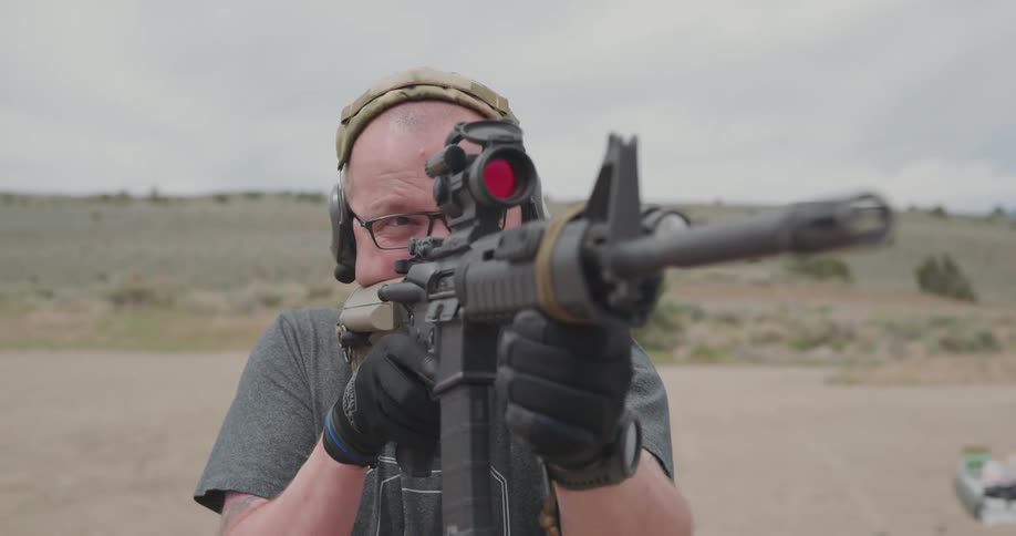 A man with glasses and gloves aiming and shooting a rifle with the sky and clouds in the background