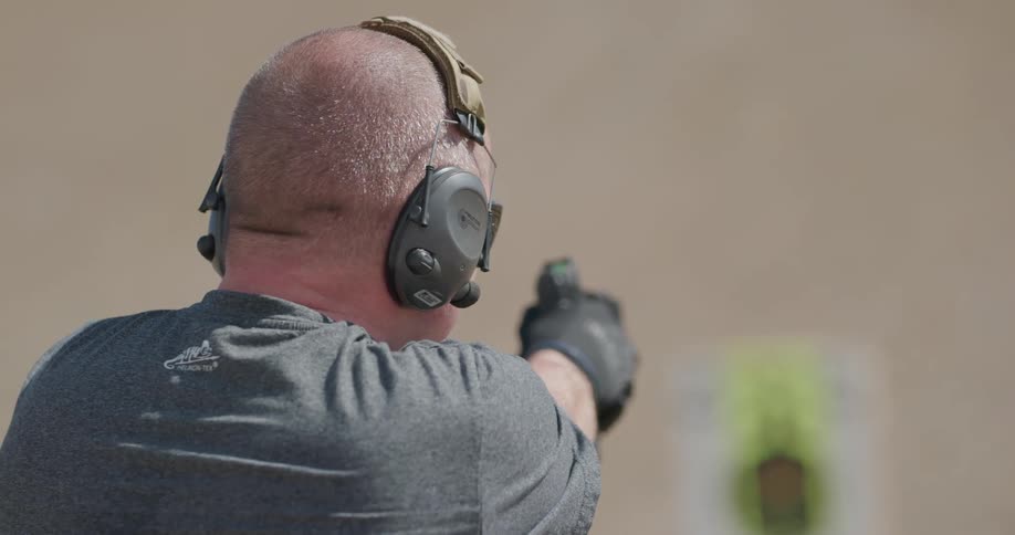 A man wearing safety earmuffs and gloves practices shooting at a paper target outdoors