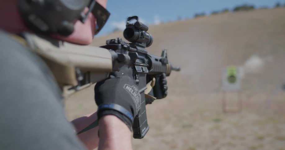A man fires a rifle at a target in field below a hill on a sunny day