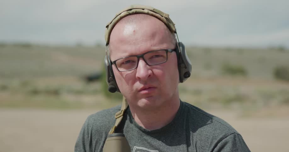 Bald man on an outdoors shooting range points an assault rifle to the camera and looks through the sight