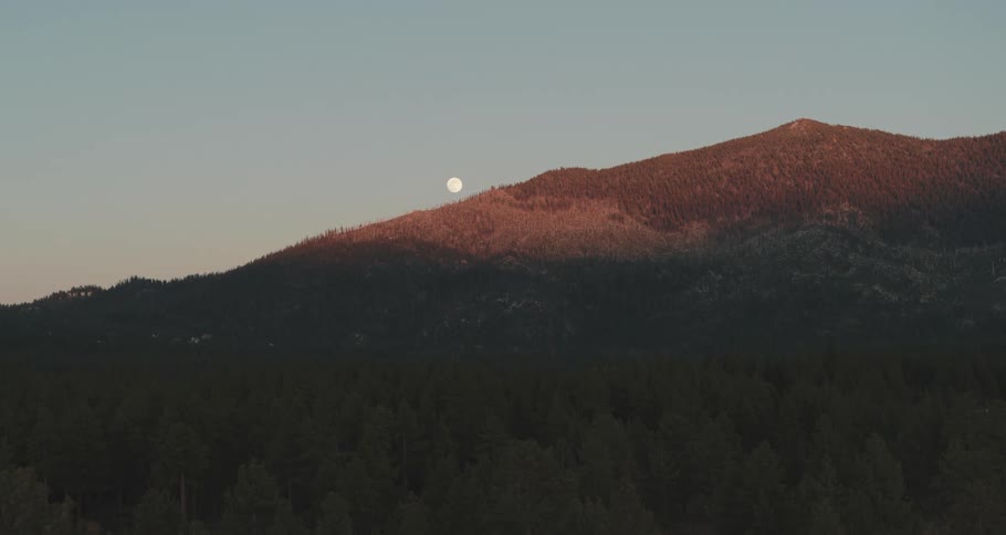 The moon peaks from behind a mountain looming over a forest in the evening