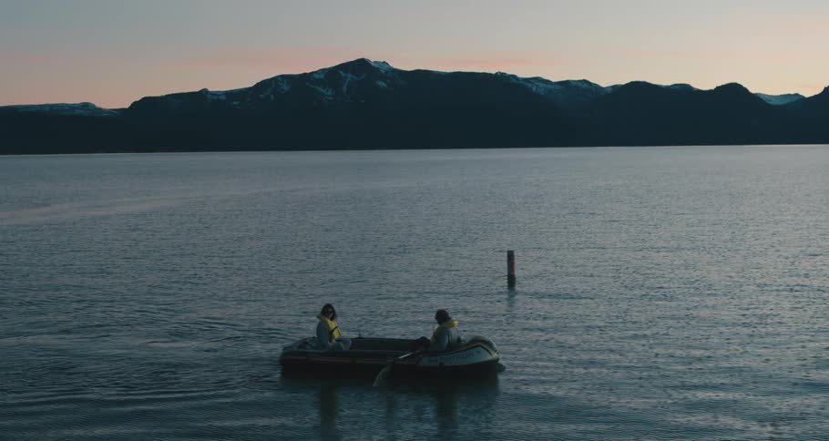 A man and woman in life jackets sit together in an inflated raft as the man rows the oars