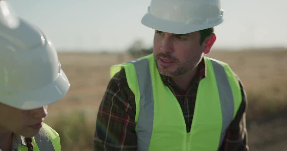 Two men and a woman with hard hats and vests standing behind a pickup truck and looking at a blueprint with the sky and a grassy field in the background