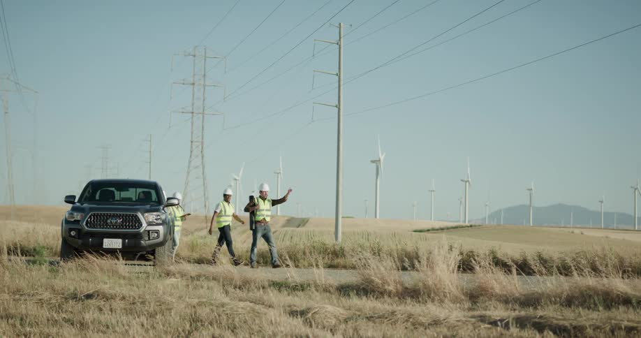 Three construction workers in yellow vests get out of a dark pickup truck to observe a site in the countryside with tall weeds and wind turbines
