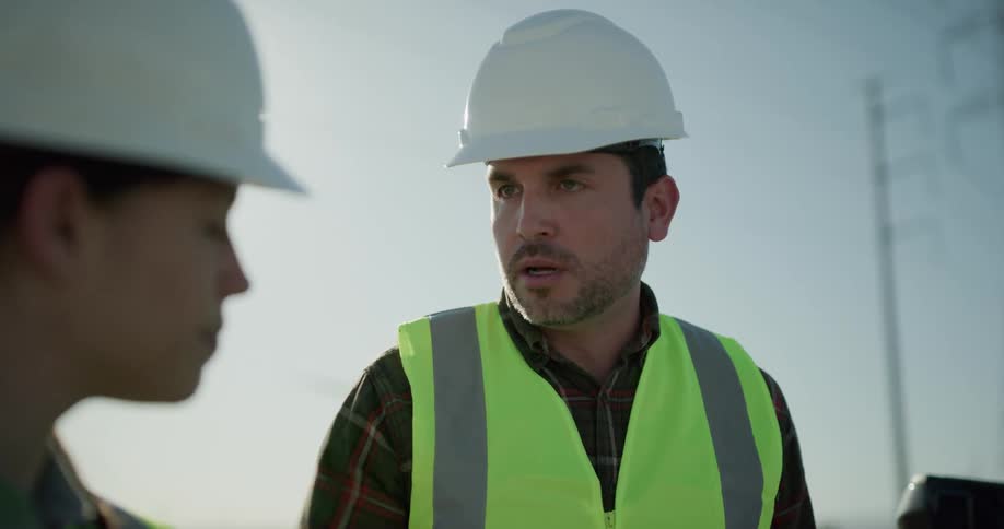 A man and a woman with hard hats and vests standing behind a pickup truck with the sky and wind turbines in the background