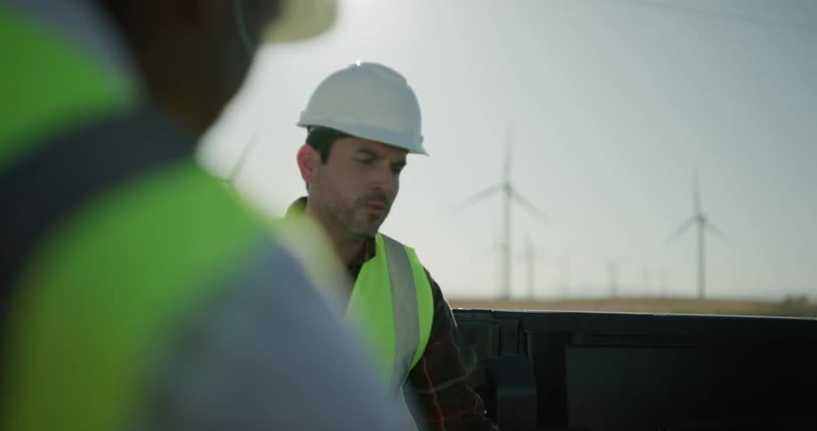 Two construction workers talk while standing by their vehicle in a field near wind turbines