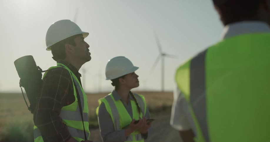 Three construction workers surveil a site on a road road running through a field filled with wind turbines
